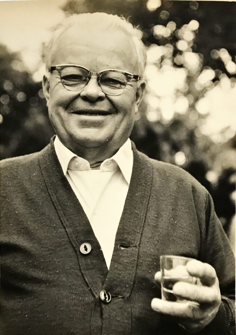 Grandpop George smiling in a cardigan, holding a shot glass
